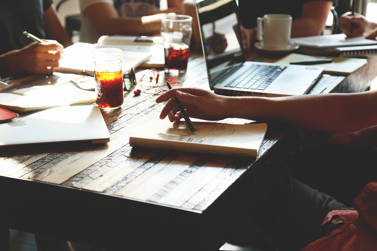Hero image — hands writing in a notebook at a busy work table, laptop and coffee nearby