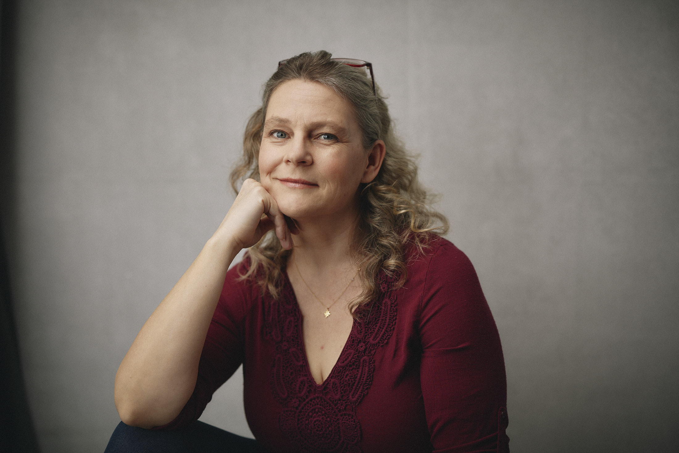 Blond woman in a burgundy casual top in front of a grey background with glasses on her head smiling into the camera